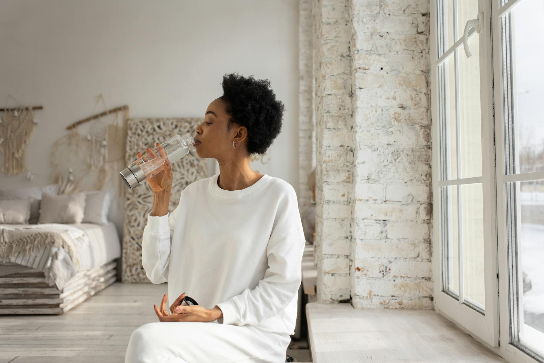 woman sitting in bedroom by window and drinking water from carafe